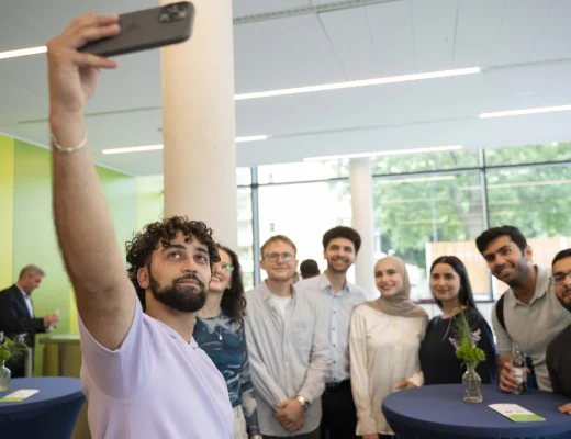 Eine Gruppe von jungen Menschen steht um einen Tisch und einer junger Mann macht ein Selfie mit dem Handy