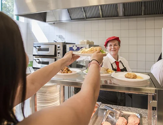 Eine Frau reicht einen Teller über eine Glastheke an eine Frau, die mit dem Rücken zur Kamera steht