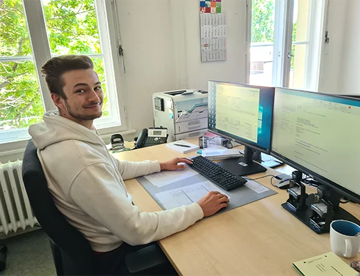 Ein Mann sitzt am Schreibtisch vor dem PC und wendet sich der Kamera zu