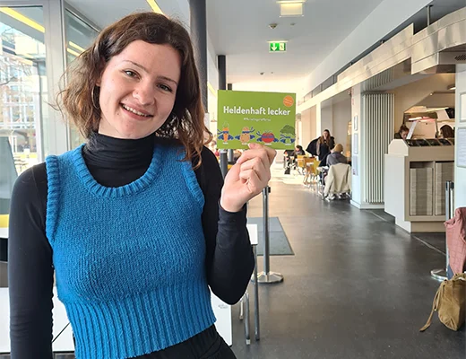 Eine junge Frau hält eine Postkarte hoch und lächelt in die Kamera