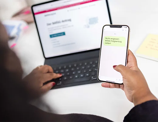 Eine Frau sitzt mit dem Rücken zur Kamera, mit dem aufgeklappten Laptop und einem Smartphone in der rechten Hand. 