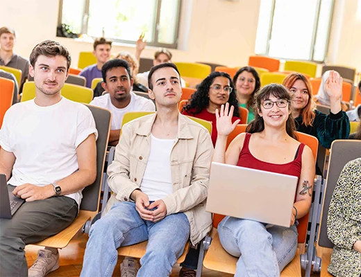 Eine Gruppe junger Menschen sitzt in einem Hörsaal, in der ersten Reihe hebt eine junge Frau mit aufgeklapptem Laptop die Hand