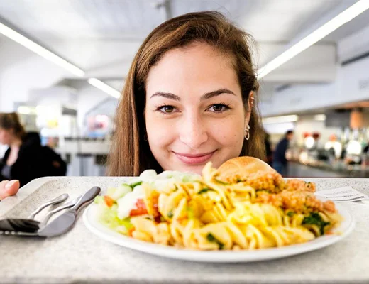 eine junge Frau mit blickt in die Kamera, in den Händen hält sie ein mit einem Teller volles Mensatablett