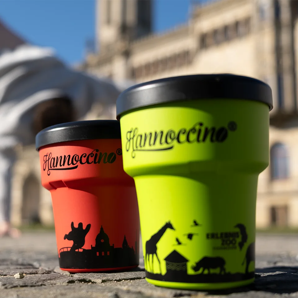 Hannoccino Mehrwegbecher in Rot und Grün mit Hannover- und Zoo-Motiv vor historischer Kulisse