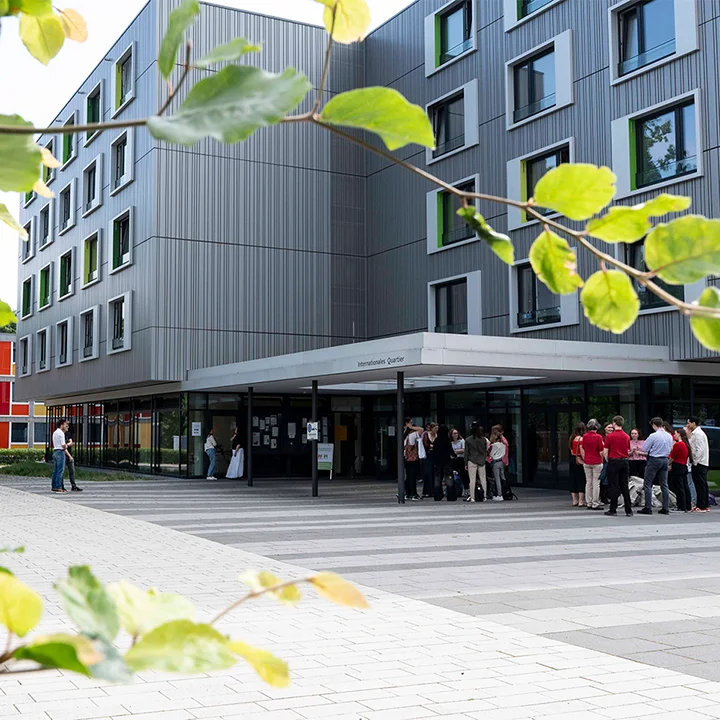 Ein Studentenwohnheim mit grauer Fassade und Menschen davor im Sonnenschein