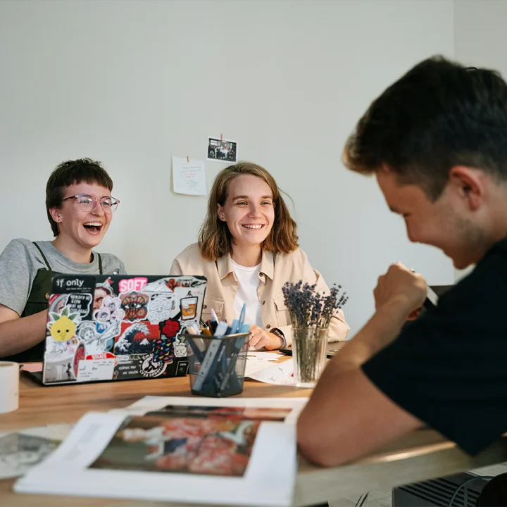Drei junge Menschen sitzen gemeinsam am Küchentisch. Einer mit dem Rücken zur Kamera, zwei im gegenüber, mit aufgeklapptem Laptop