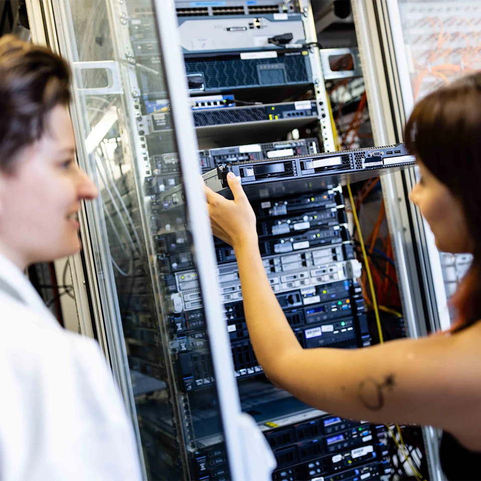 Zwei Personen stehen vor einer Serveranlage mit dem Rücken zur Kamera, eine entnimmt eine Festplatte