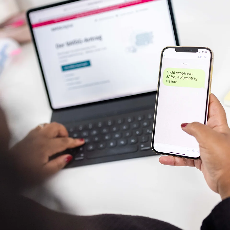 Eine Frau sitzt mit dem Rücken zur Kamera, mit dem aufgeklappten Laptop und einem Smartphone in der rechten Hand. 