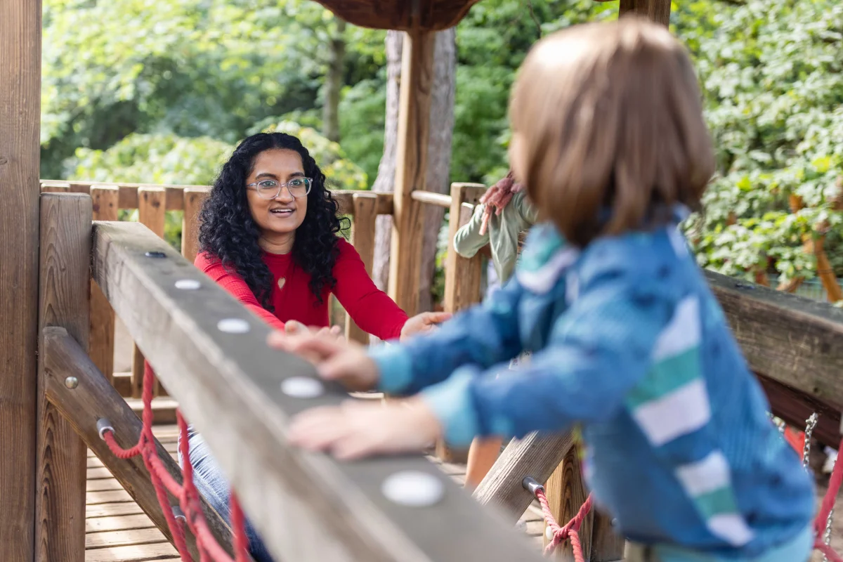 Eine junge Frau mit dunklen Haaren ist mit einem kleinem Kind auf einer Holzburg auf dem Spielplatz