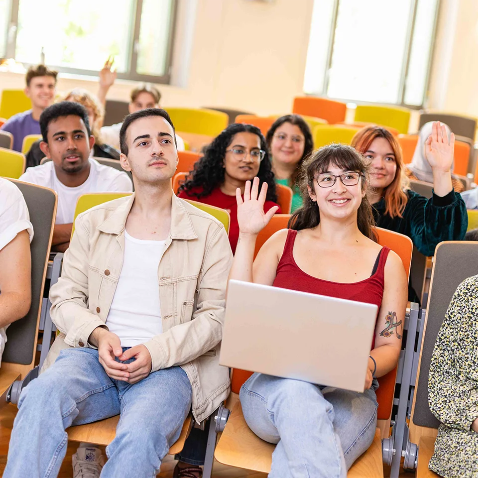 eine Gruppe von Personen sitzt in einem Hörsaal, eine junge Frau hebt die Hand