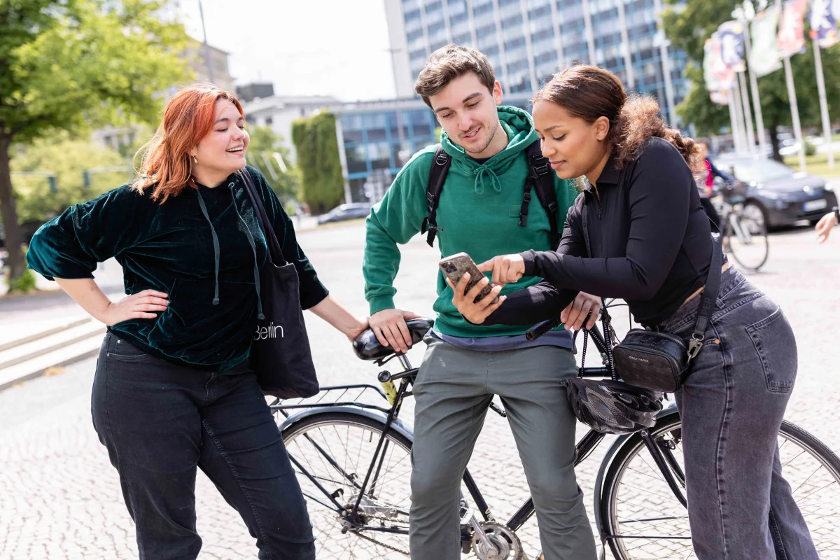 Zwei Studentinnen unterhalten sich mit einem Student, der an einem Fahrrad lehnt