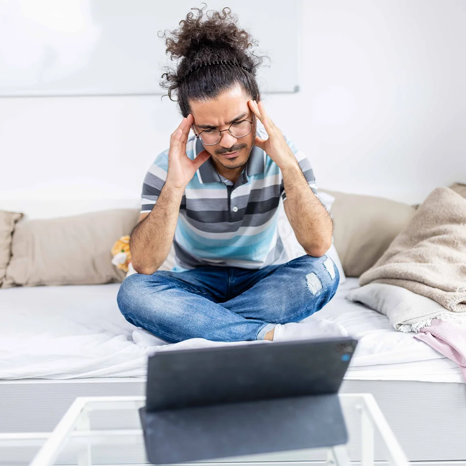 Ein junger Mann sitzt im Schneidersatz auf dem Bett und schaut verzweifelt auf einen Laptop-Bildschirm